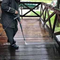 Mężczyzna w ciemnej kurtce i gumowych butach czyści drewniany taras za pomocą myjki ciśnieniowej, widoczny efekt czyszczenia i brudna woda.