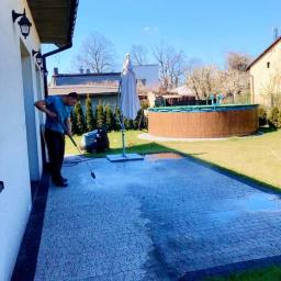 ZaCLEANacz Żdzary - Mężczyzna czyści kostkę brukową myjką ciśnieniową na tle ogrodu z basenem i parasolem.