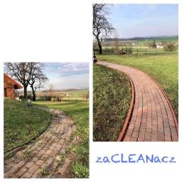 ZaCLEANacz Żdzary - Porównanie stanu kostki brukowej przed i po czyszczeniu myjką ciśnieniową, widoczne dwie ścieżki z kostki, jedna porośnięta mchem i chwastami, druga czysta, na tle zielonego trawnika i krajobrazu...