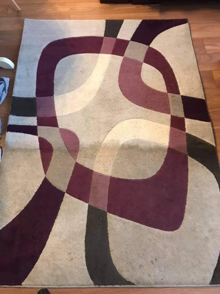 Dywan z geometrycznym wzorem w odcieniach beżu, brązu i fioletu, leżący na drewnianej podłodze. Widoczne plamy sugerują potrzebę czyszczenia.