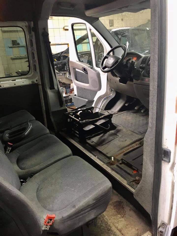 Wnętrze furgonetki w trakcie demontażu, widoczne wymontowane elementy deski rozdzielczej i podłogi, szare fotele z pomarańczowymi zatrzaskami, otwarte drzwi kabiny.
