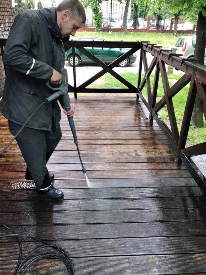 Mężczyzna w ciemnej kurtce i gumowych butach czyści drewniany taras za pomocą myjki ciśnieniowej, widoczny efekt czyszczenia i brudna woda.