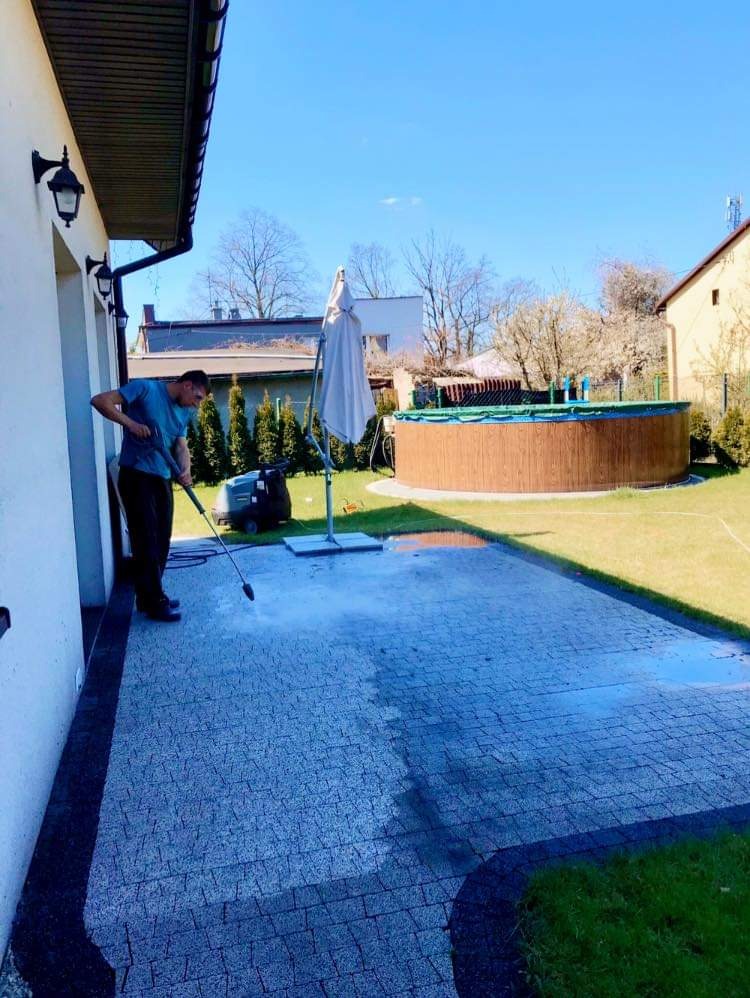 Mężczyzna czyści kostkę brukową myjką ciśnieniową na tle ogrodu z basenem i parasolem.