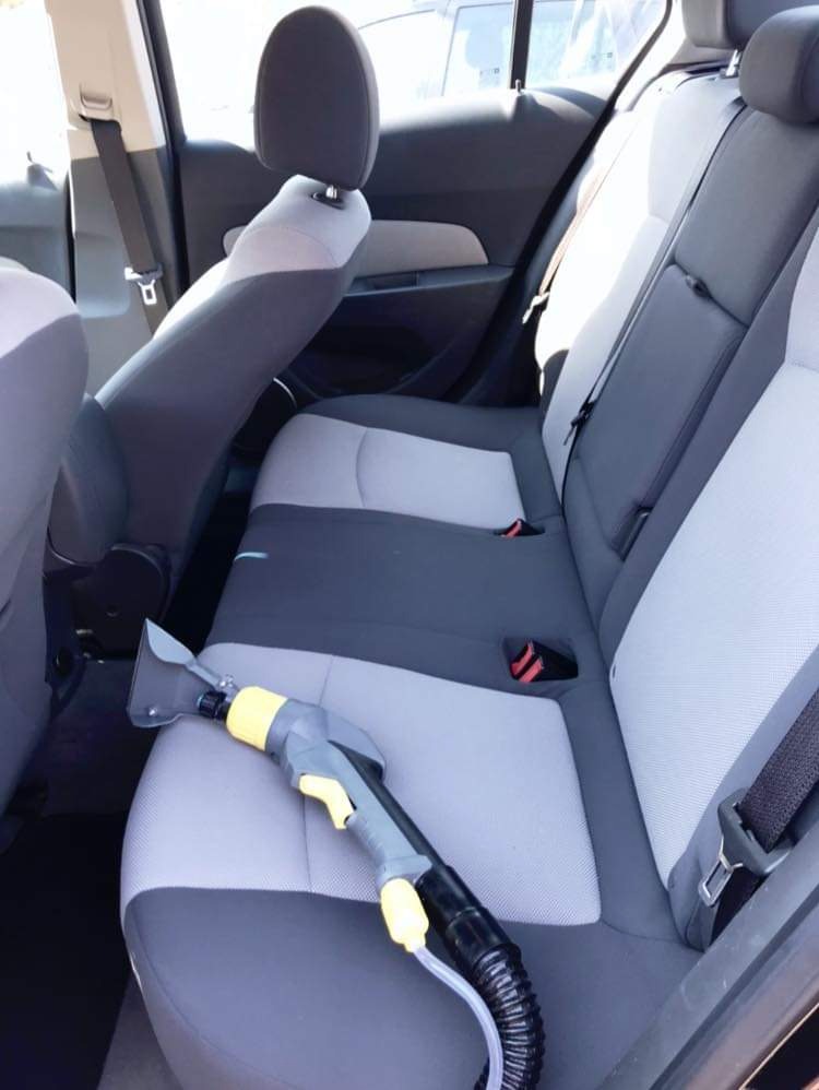 Wnętrze samochodu z szarymi fotelami, na tylnym siedzeniu leży odkurzacz piorący z żółtymi elementami, przygotowany do czyszczenia tapicerki.