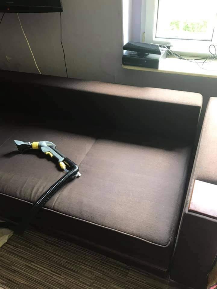 Brązowa sofa z dyszą czyszczącą na siedzisku, gotowa do rozpoczęcia procesu czyszczenia tapicerki. W tle widoczne okno i sprzęt elektroniczny.