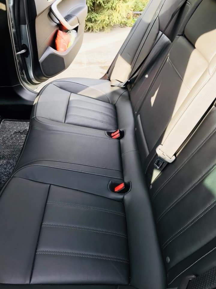 Czarne, skórzane tylne siedzenia samochodu po czyszczeniu, z widocznymi szwami i elementami pasów bezpieczeństwa. W tle otwarte drzwi i zielona roślinność.