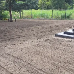 Przygotowany teren pod trawnik z widocznymi śladami po maszynach, z elementami architektury ogrodowej w tle: schody z kostki brukowej, donica i zieleń.