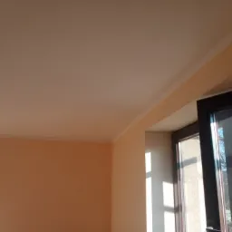 Pomalowane na brzoskwiniowo wnętrze pokoju z białym sufitem, widok na okno z ciemną ramą i padające promienie słoneczne.