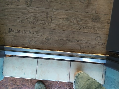 Połączenie podłogi z paneli imitujących drewno z płytkami ceramicznymi, oddzielone metalową listwą progową, widok z góry, fragment nogi w jeansach.