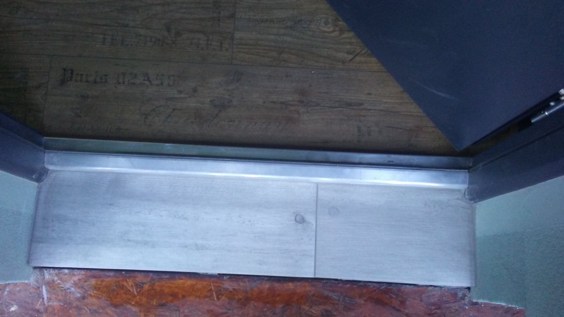 Połączenie dwóch rodzajów podłóg: ciemnej drewnopodobnej z nadrukiem 'Paris 02ASS' i jasnej, z metalową listwą progową, widoczne ślady po remoncie.