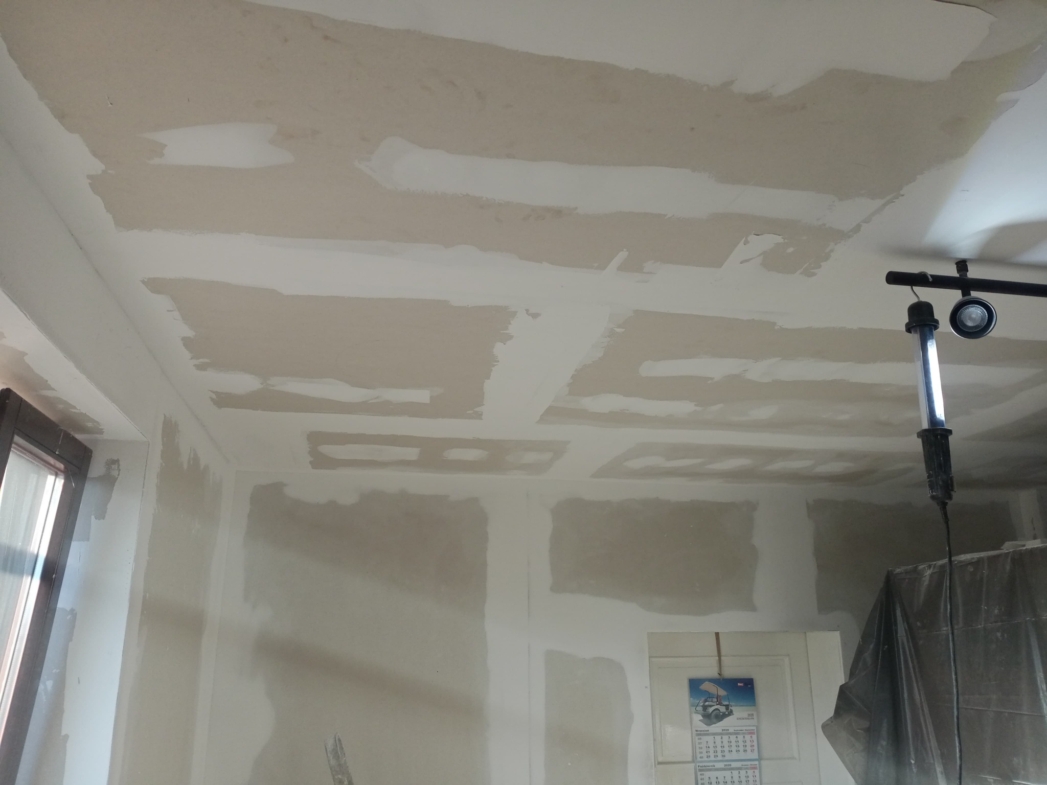 Pokój w trakcie prac wykończeniowych: ściany i sufit pokryte szpachlą, widoczne ślady po łatach, lampa robocza oświetla pomieszczenie, kalendarz wisi na drzwiach.