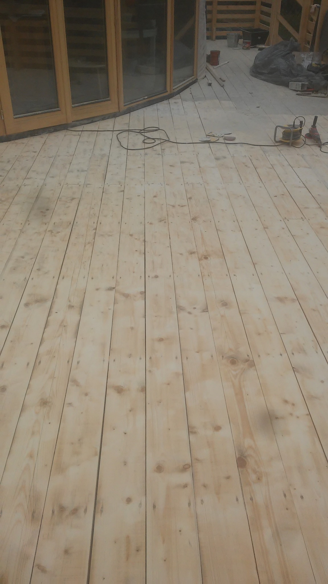 Drewniana podłoga tarasu w trakcie budowy, widoczne narzędzia i maszyny do obróbki drewna, okna tarasowe w tle.