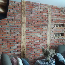 Ściana z czerwonej cegły z drewnianymi belkami i wnękami z świecami, widok na fragment schodów i część umeblowanego salonu.