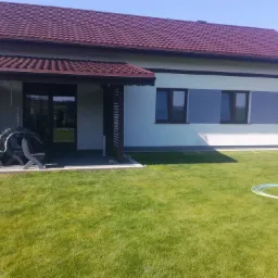 Nowoczesny dom jednorodzinny z brązowym dachem i jasną elewacją, widoczne trzy szare pasy na ścianie pomiędzy oknami, zadbany trawnik z rozłożonym wężem ogrodowym.