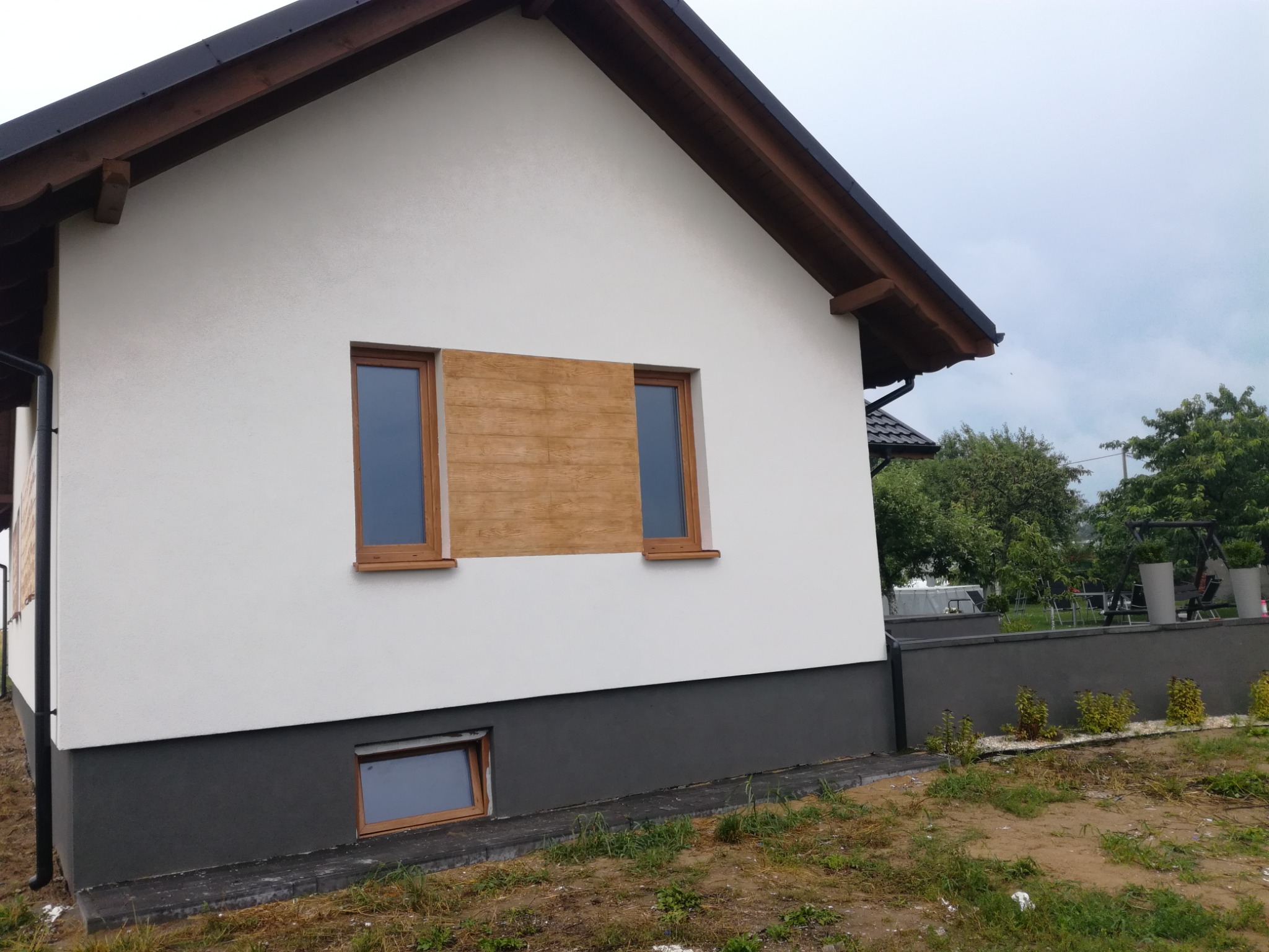 Boczna ściana domu z białą elewacją i ciemnoszarą podmurówką, z dwoma oknami obok drewnianej, dekoracyjnej wstawki imitującej drewno, okno w podmurówce, widoczny fragment dachu i rynny, trawa...