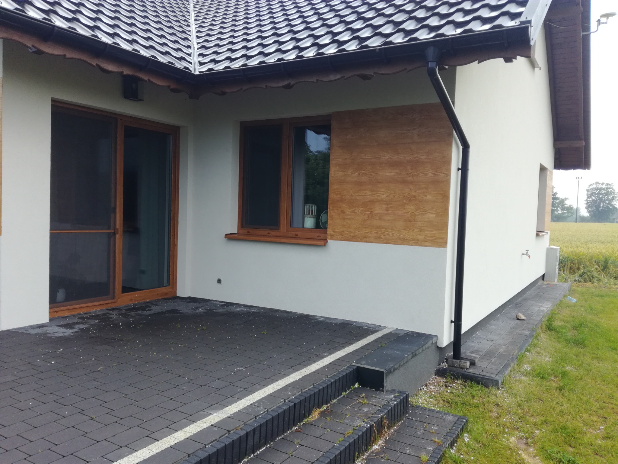 Wykończona elewacja domu jednorodzinnego z drewnopodobną okładziną, widoczne okna z drewnianą ramą, czarna rynna, szara kostka brukowa na tarasie i schodach.