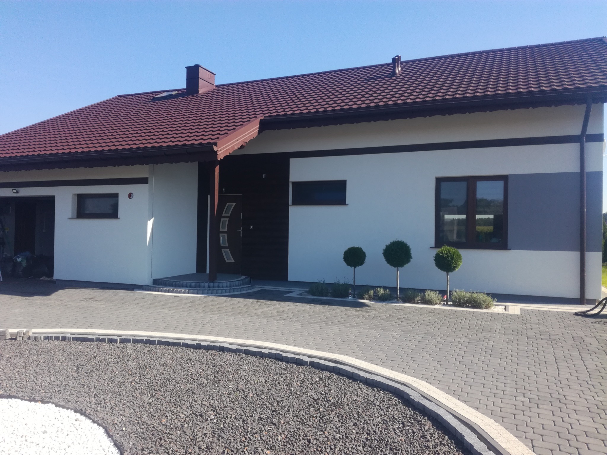 Nowoczesny dom jednorodzinny z brązowym dachem i jasną elewacją, z trzema kulistymi drzewkami ozdobnymi przed oknem i brukowanym podjazdem z kamieniami ozdobnymi.