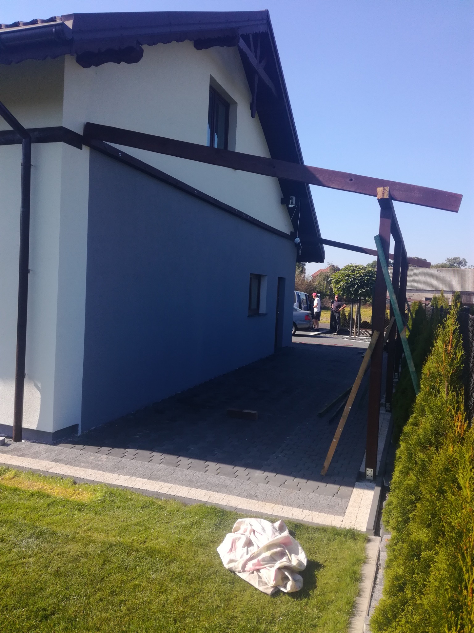 Świeżo pomalowana elewacja domu w odcieniach szarości z elementami drewnianej konstrukcji zadaszenia w trakcie budowy, widoczne tuje i trawnik.