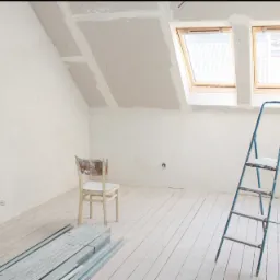 Poddasze w trakcie wykańczania płytami kartonowo-gipsowymi, widoczne łączenia szpachlowane, drewniana podłoga pomalowana na biało, metalowe profile na podłodze, drabina malarska i krzesło.