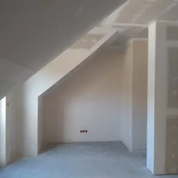 Poddasze w trakcie wykańczania: ściany i sufit pokryte płytami gipsowo-kartonowymi, widoczne łączenia przygotowane do szpachlowania, trzy gniazdka elektryczne na ścianie, surowy beton na podłodze.