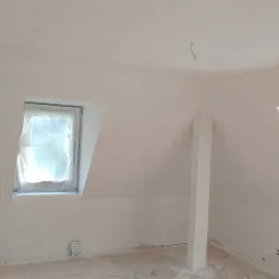 Pokój na poddaszu po świeżym pomalowaniu na biało, okno zabezpieczone folią, podłoga zakryta materiałem ochronnym, widoczne ślady szpachli na ścianach.