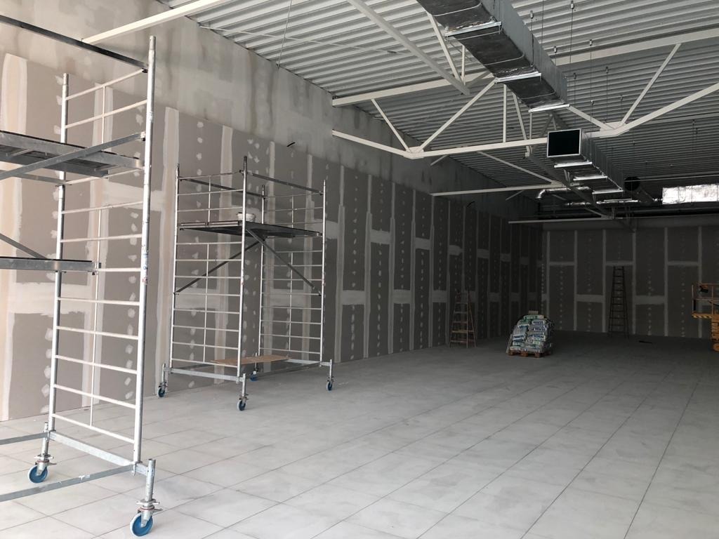 Wnętrze w trakcie prac wykończeniowych: ściany z zamontowaną płytą kartonowo-gipsową, widoczne rusztowania na kółkach i worki z materiałami budowlanymi w tle, surowy stan wykończenia.
