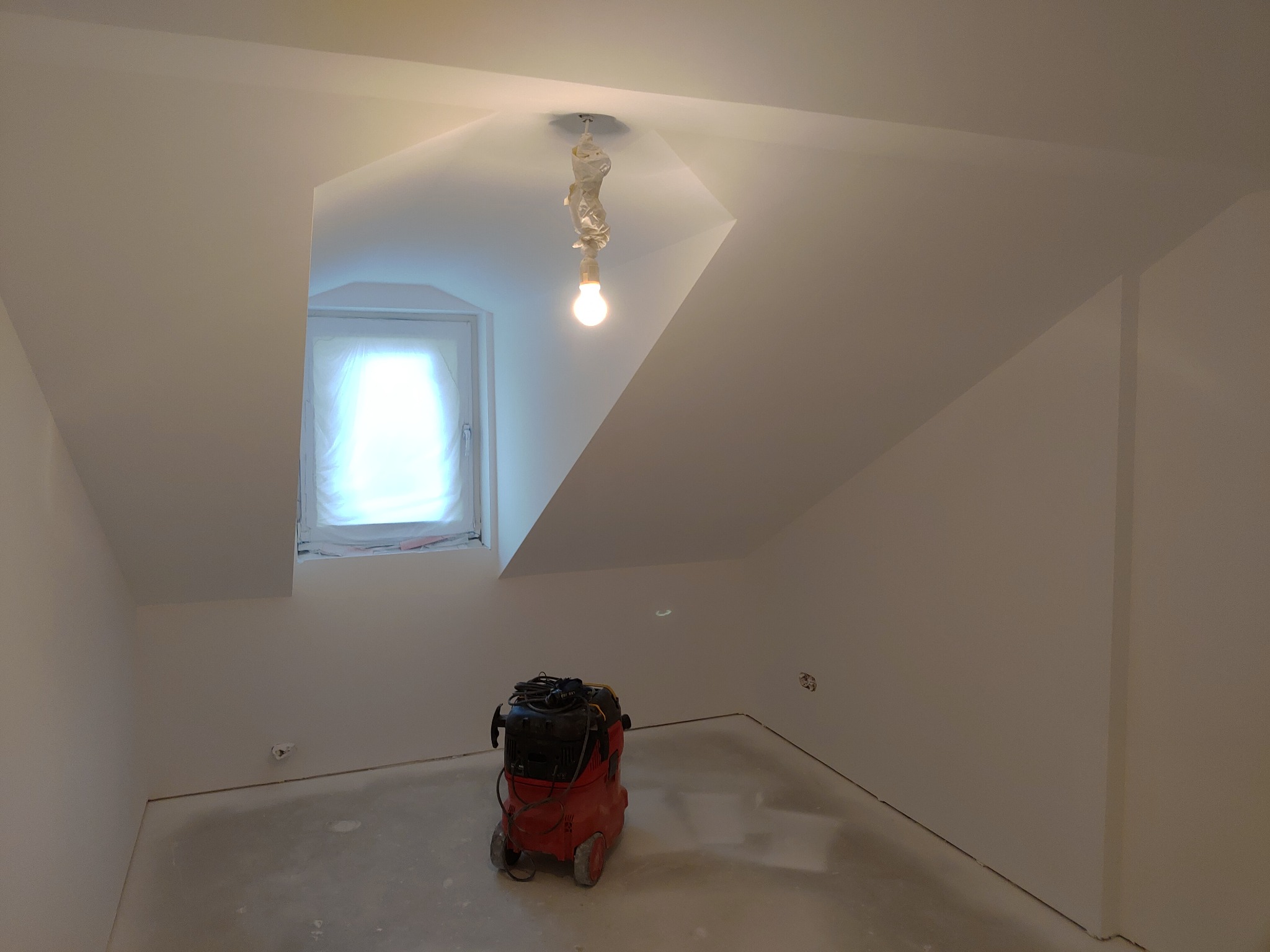Poddasze po szpachlowaniu i malowaniu, z zabezpieczonym oknem i lampą, widoczny odkurzacz przemysłowy na zaszpachlowanej podłodze.