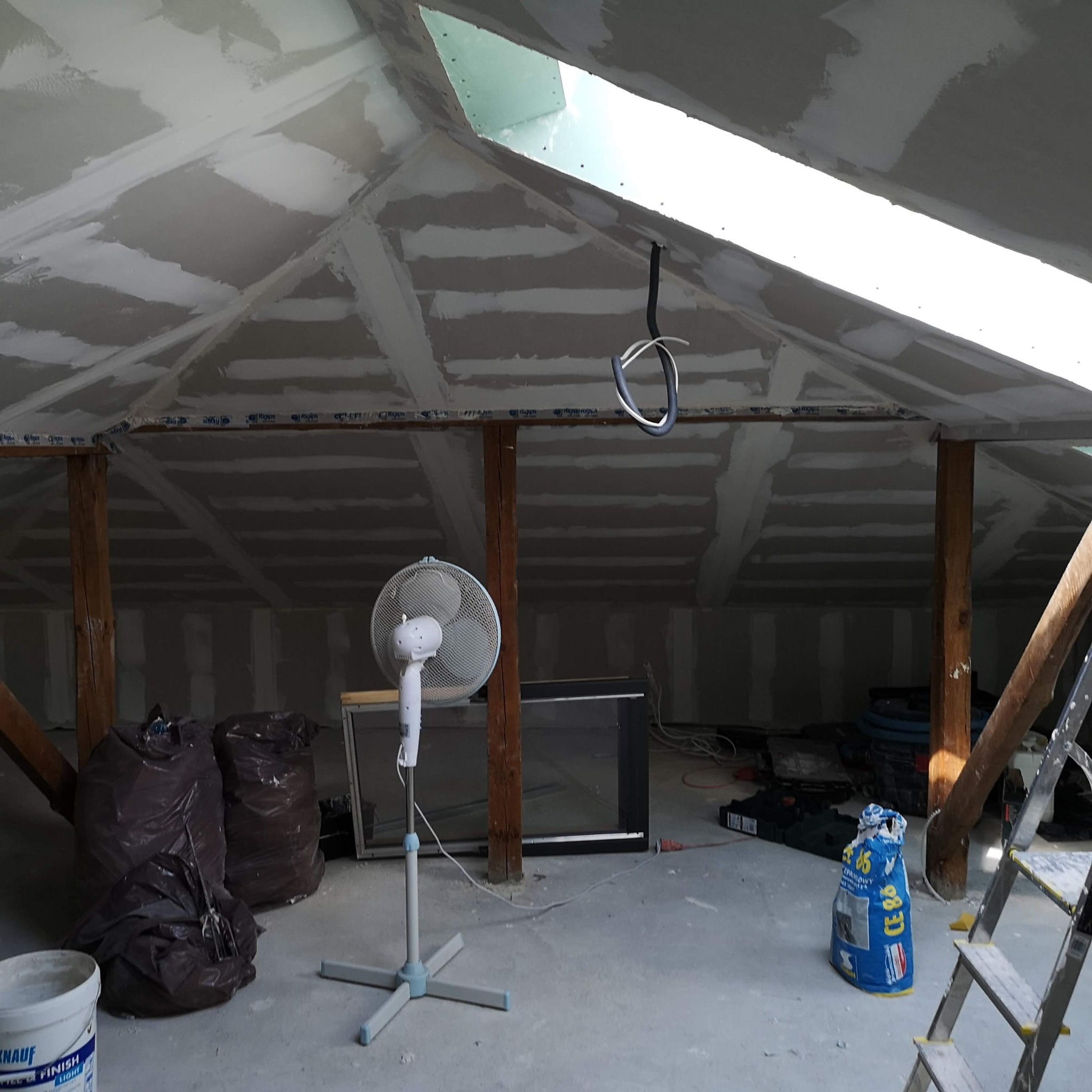 Poddasze w trakcie prac wykończeniowych: widoczne płyty kartonowo-gipsowe na skośnych ścianach i suficie, częściowo pokryte masą szpachlową, drewniane belki konstrukcyjne, wentylator stojący, worki...