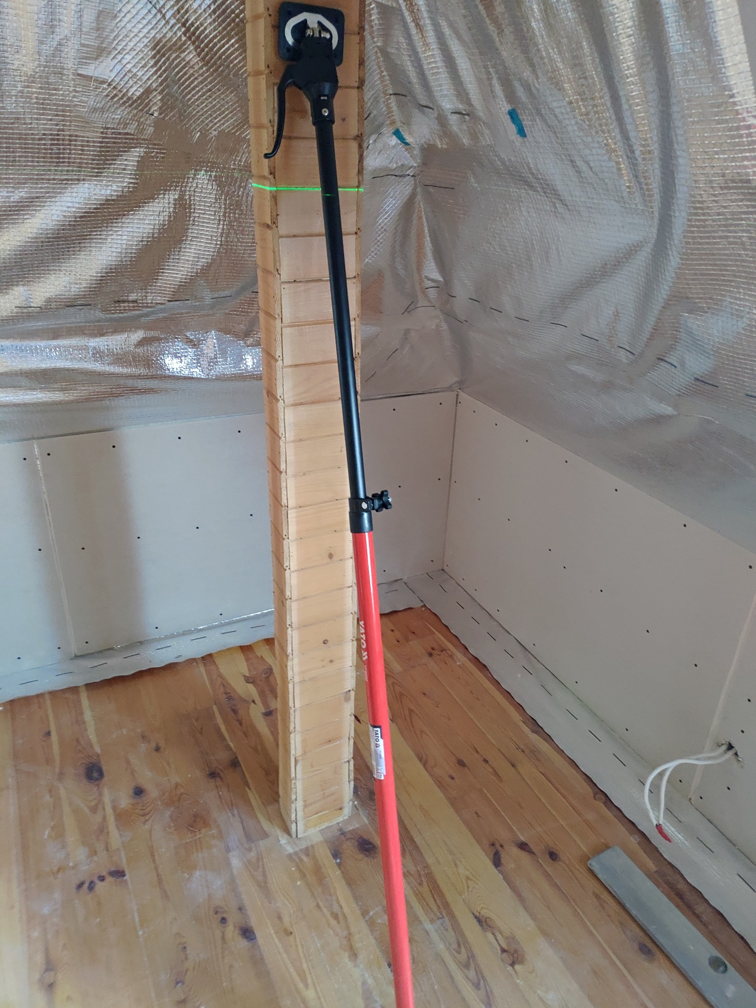 Poddasze w trakcie izolacji: widoczna folia termoizolacyjna, zamontowane płyty gipsowo-kartonowe, drewniana konstrukcja wzmacniająca oraz narzędzie do mocowania płyt. Na podłodze widoczne ślady...