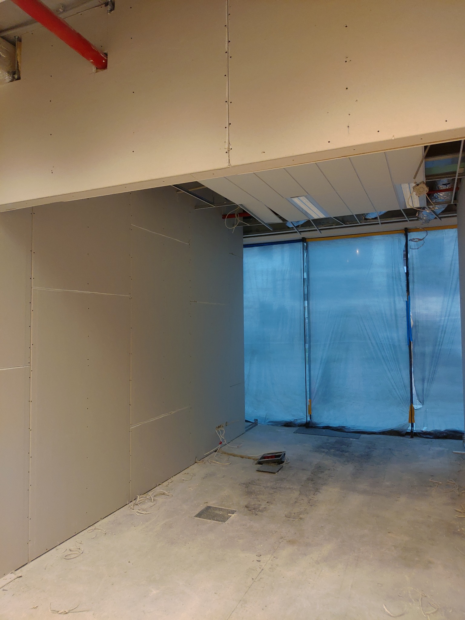 Montaż płyt karton gips na ścianie w pomieszczeniu z widocznymi przewodami elektrycznymi i zabezpieczeniem folią malarską na oknach.