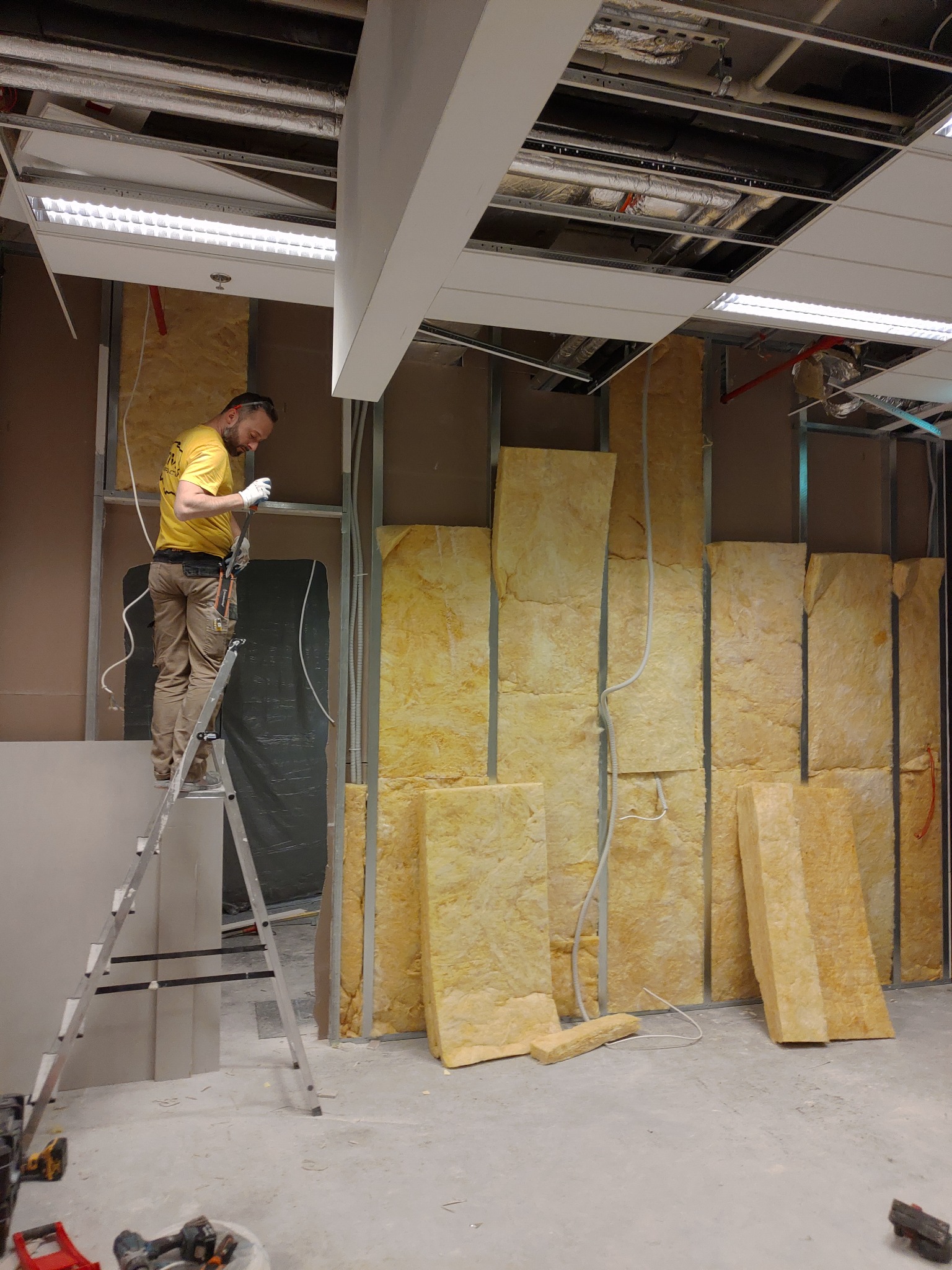 Pracownik na drabinie montuje izolację z wełny mineralnej w metalowym stelażu ściany działowej w pomieszczeniu z odsłoniętą instalacją pod sufitem, widoczne narzędzia i płyta gipsowo-kartonowa.