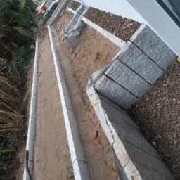 Budowa obrzeży rabaty z kostki granitowej, widoczny wykop, piasek i kamień ozdobny.