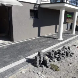 Ułożona ścieżka z szarej kostki brukowej z białymi akcentami prowadząca do domu, obok sterta kostki przygotowana do dalszych prac.