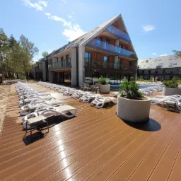 Nowoczesny hotel z basenem zewnętrznym w Łebie, widok na drewniany taras z leżakami i plac budowy z koparką w tle.