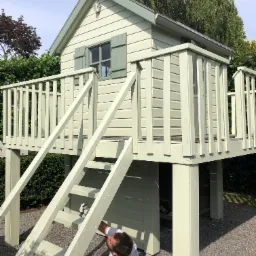 Mężczyzna w białym ubraniu maluje pędzlem na drabinie drewniany domek dla dzieci w odcieniach zieleni, domek na podwyższeniu z tarasem i schodami, otoczony zielenią i żwirowym podłożem.