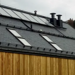 Ciemnoszary dach z dachówki łupkowej z oknami dachowymi, kominami i panelami słonecznymi; drewniana elewacja budynku w dolnej części kadru.