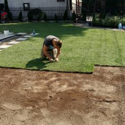 Maciej Żychowski - Mężczyzna na kolanach dopasowuje brzeg rolki trawy do przygotowanego podłoża w ogrodzie, w tle widoczny fragment ułożonej trawy, dom i ogrodzenie.
