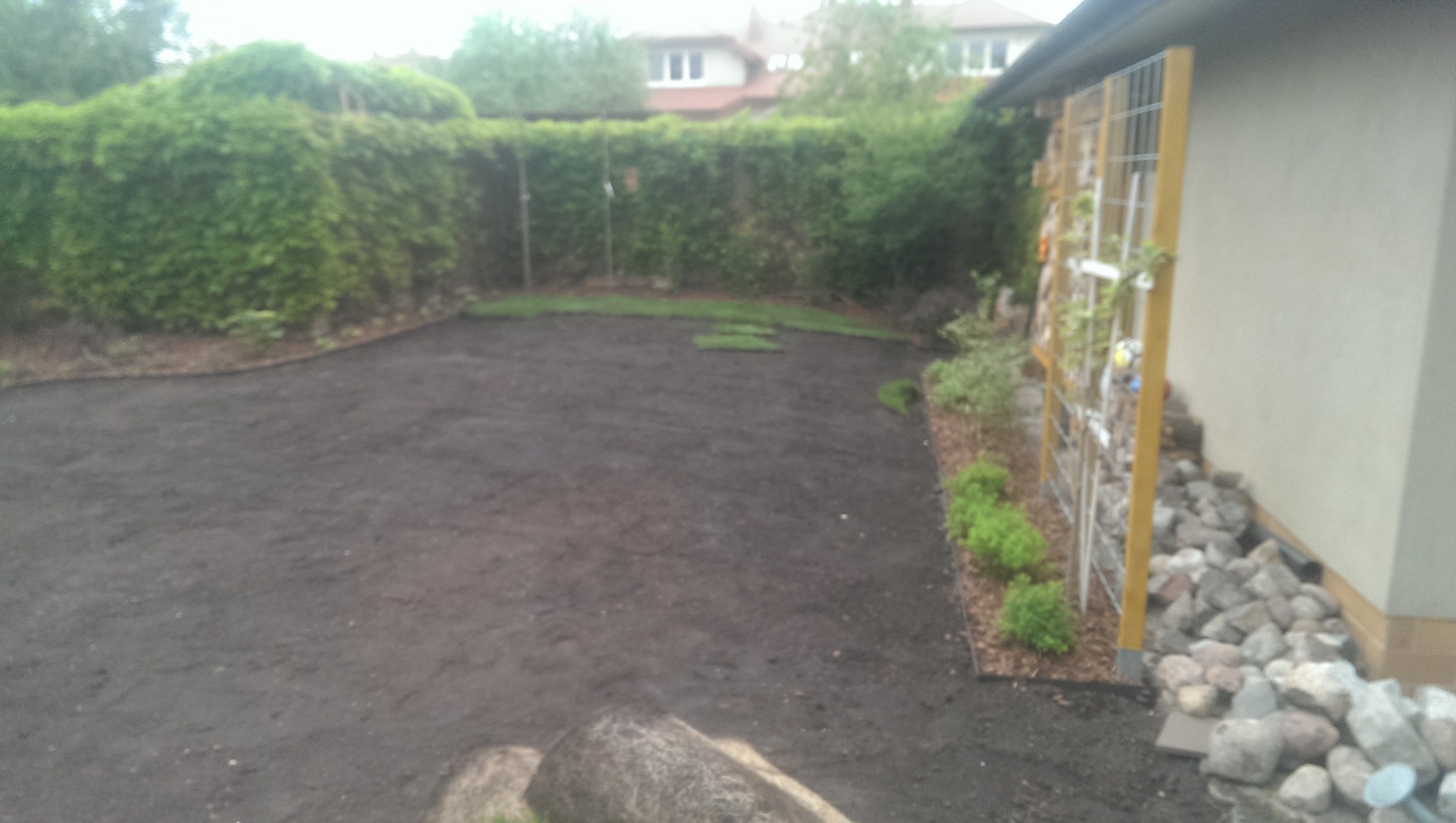 Przygotowany teren pod trawnik z rolki, widoczne fragmenty rozłożonej darni, obrzeża z kory i kamieni, oraz konstrukcja do pnących roślin przy ścianie budynku.