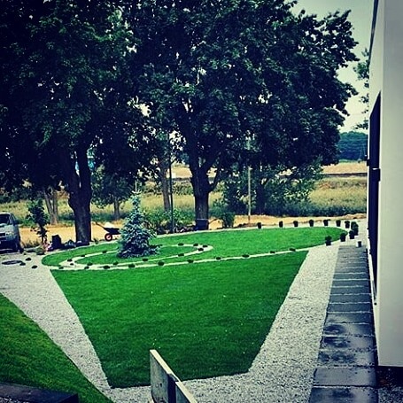 Nowoczesny ogród z trawnikiem, ścieżkami z jasnego żwiru i ozdobnymi nasadzeniami w doniczkach, widok z boku domu, w tle pole i drzewa.