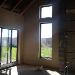 Wnętrze surowego domu po nałożeniu gładzi, widoczne okna z widokiem na zewnątrz, wiaderka i węże na podłodze, oraz konstrukcja rusztowania przy ścianie z cegieł.