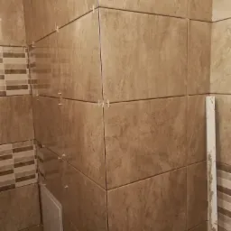 Brązowe płytki ścienne imitujące kamień, ułożone z użyciem krzyżyków dystansowych, widoczny fragment ściany z dekoracyjną mozaiką i biała listwa przypodłogowa.