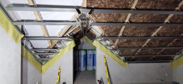 Montaż izolacji z wełny mineralnej na poddaszu, widoczne profile metalowe, folia paroizolacyjna i rolki wełny w tle.