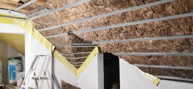 Montaż izolacji z wełny mineralnej w przestrzeni poddasza, widoczne metalowe profile i żółta folia paroizolacyjna, drabina oparta o ścianę, worki z materiałami budowlanymi w tle.