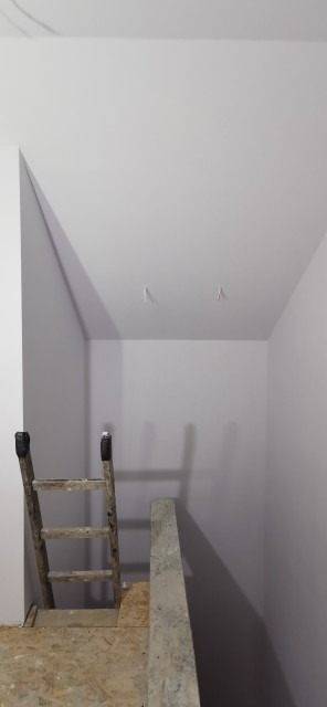 Wnętrze pomalowane na szaro z drewnianą drabiną opartą o ścianę, widoczne wystające kable ze ściany, podłoga wyłożona płytami OSB.