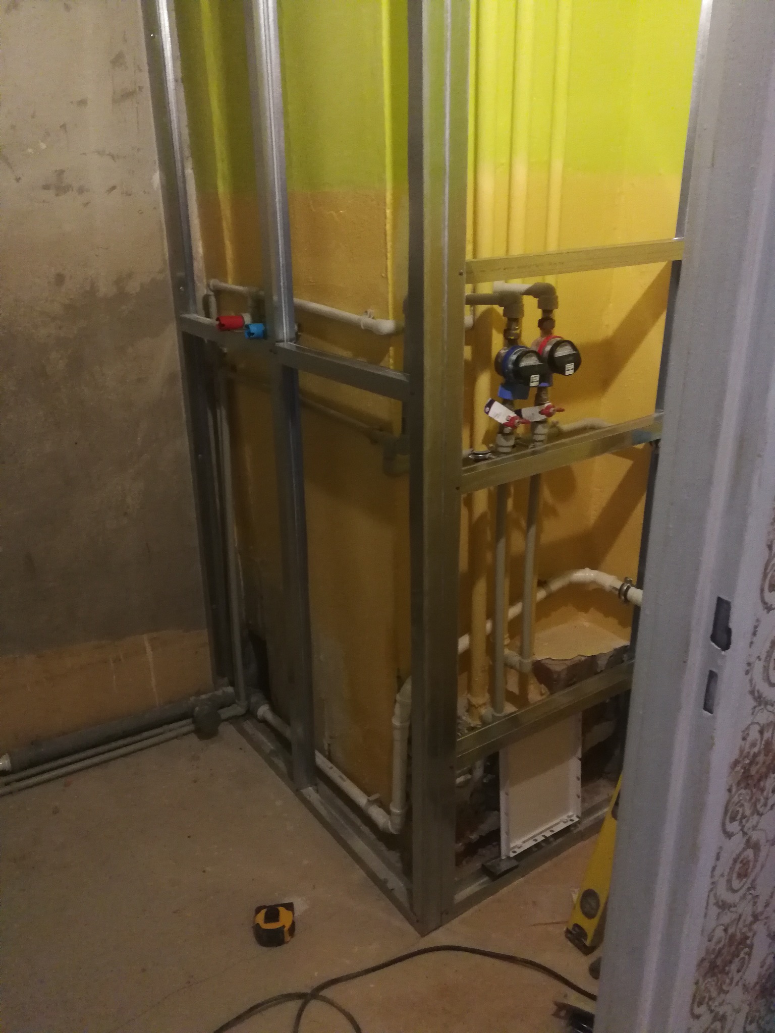 Stelaż z profili metalowych przygotowany pod zabudowę instalacji wodnej i kanalizacyjnej w trakcie prac remontowych łazienki, widoczna żółta ściana w tle, rury i zawory, na podłodze leży zwinięta...