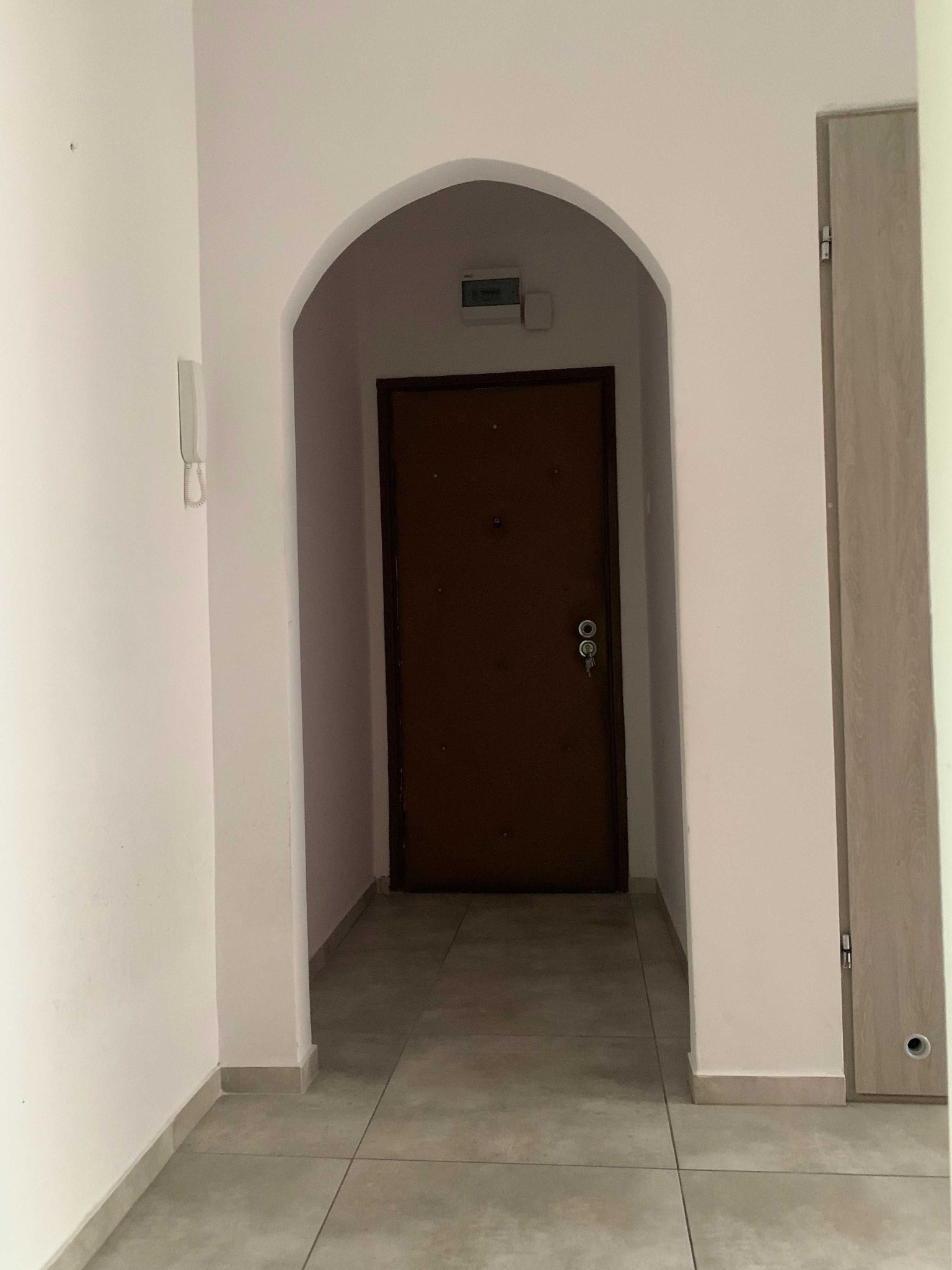 Wnętrze korytarza z łukowym przejściem do brązowych drzwi wejściowych, jasne ściany i szara podłoga.