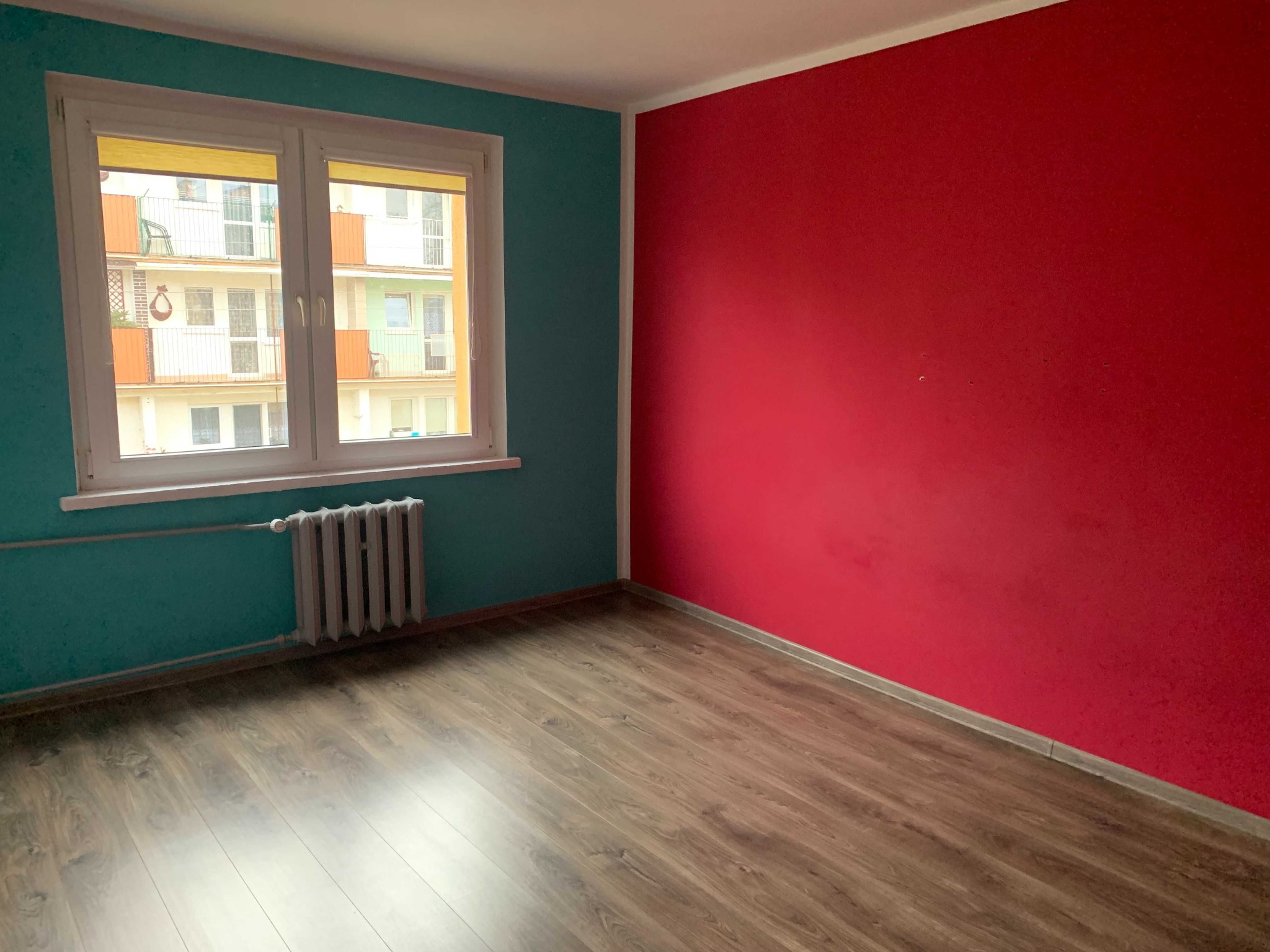Pusty pokój po odświeżeniu: turkusowa ściana z oknem i kaloryferem, czerwona ściana, panele podłogowe imitujące drewno.