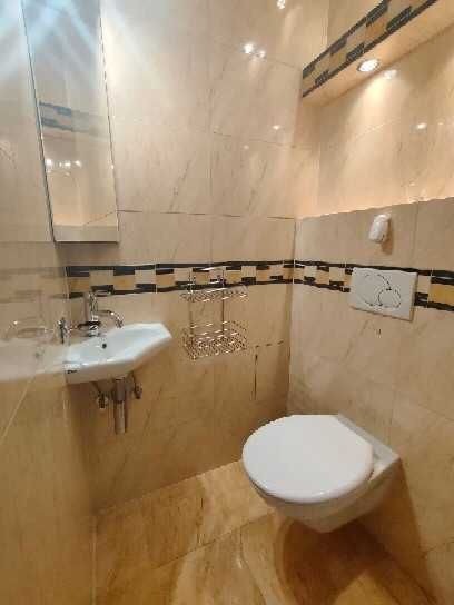 Wykończona łazienka z beżowymi płytkami imitującymi marmur, wisząca toaleta, umywalka z lustrem, metalowa półka na kosmetyki i dekoracyjny pas z mozaiki.