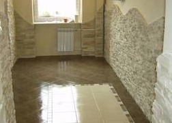 Korytarz wykończony w jasnych odcieniach beżu i brązu. Ściany częściowo pokryte dekoracyjnym kamieniem, podłoga z błyszczących płytek. Widoczne okno z grzejnikiem pod nim.