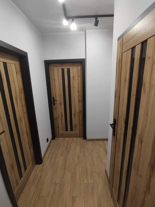 Korytarz z trzema drewnianymi drzwiami z czarnymi wstawkami, oświetlony listwą z trzema reflektorami punktowymi, podłoga wyłożona panelami imitującymi drewno.